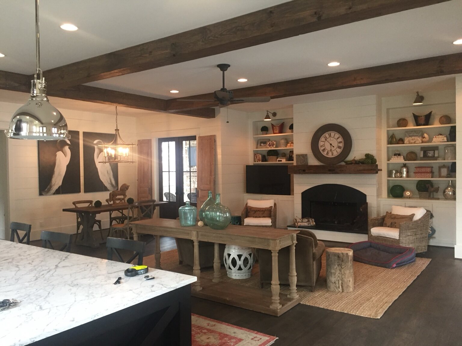 Open living room with fireplace, built-in shelving, shiplap walls, and timber beams
